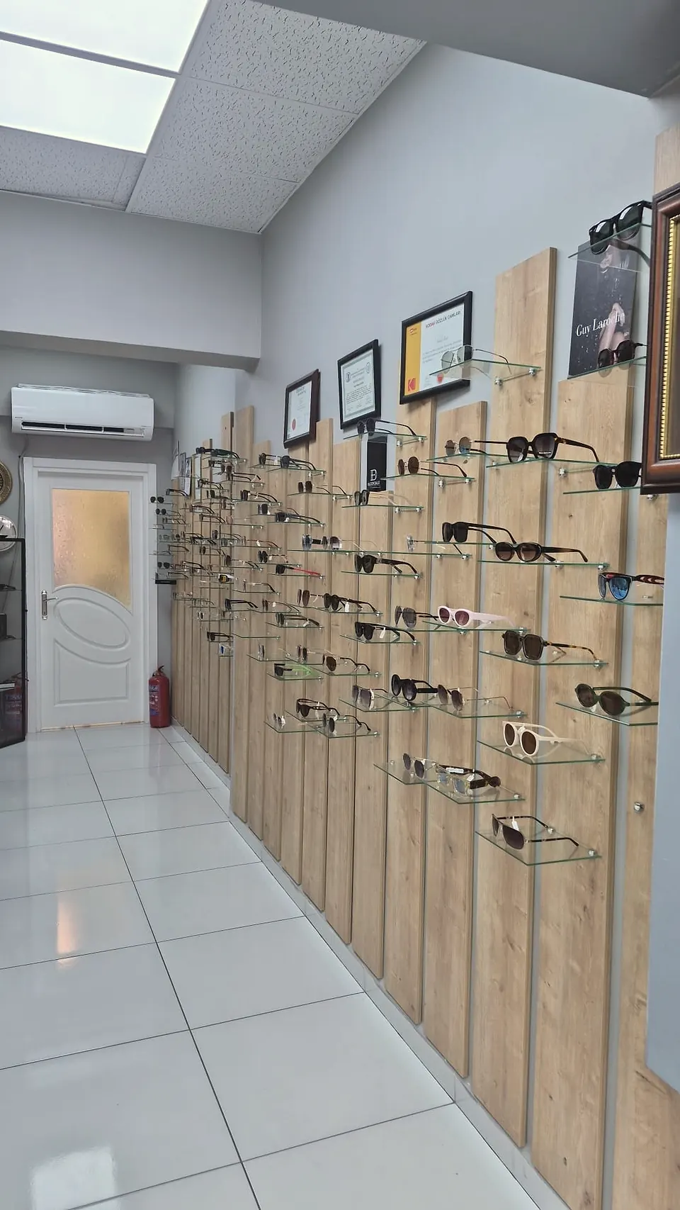 Sinan Optik corridor — wooden display panels holding a wide selection of sunglasses and prescription frames, certificates and Guy Laroche poster